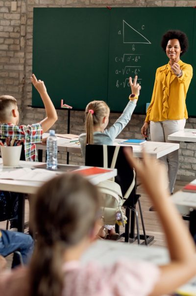 Happy black teacher pointing at his elementary students who are raising hands to answer the question during a class. Happy African American teacher holding a class and aiming at her students who are raising their hands to answer the question in the classroom.